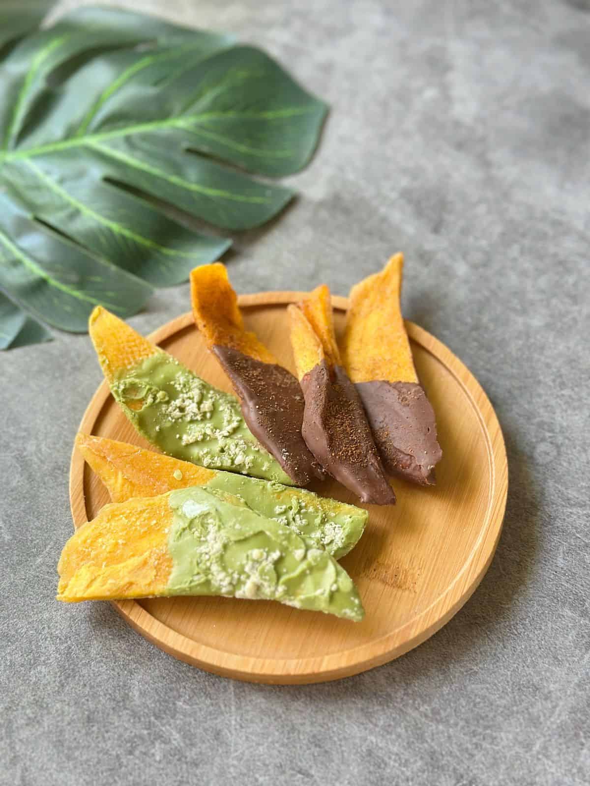 20 Vietnamesische Mitbringsel für Deine Reise Half-dipped chocolate dried mango and matcha white chocolate covered dried mango pieces arranged on a wooden plate.