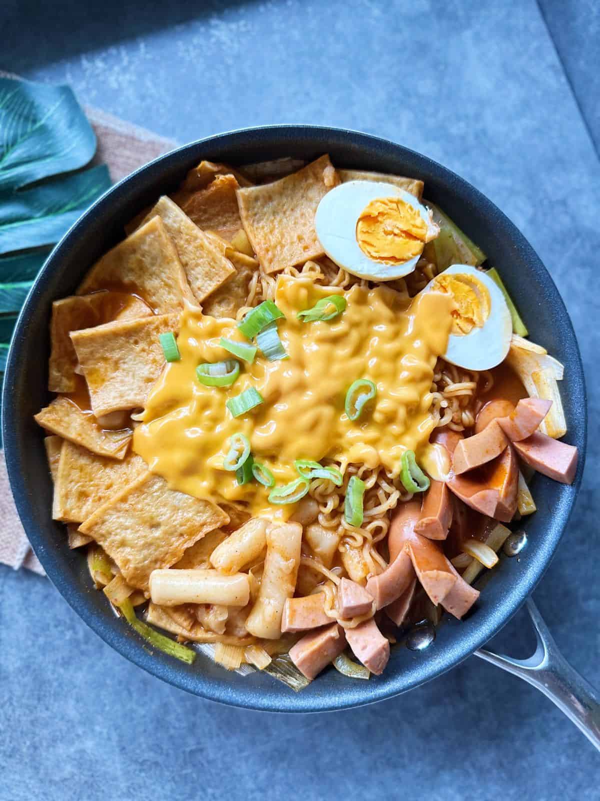 Pfanne mit würzigem Rabokki – koreanische Reiskuchen (Tteokbokki) und Ramyeon in roter Gochujang-Soße, mit Fischkuchen, Wurst, Ei, Lauch und geschmolzenem Käse garniert.