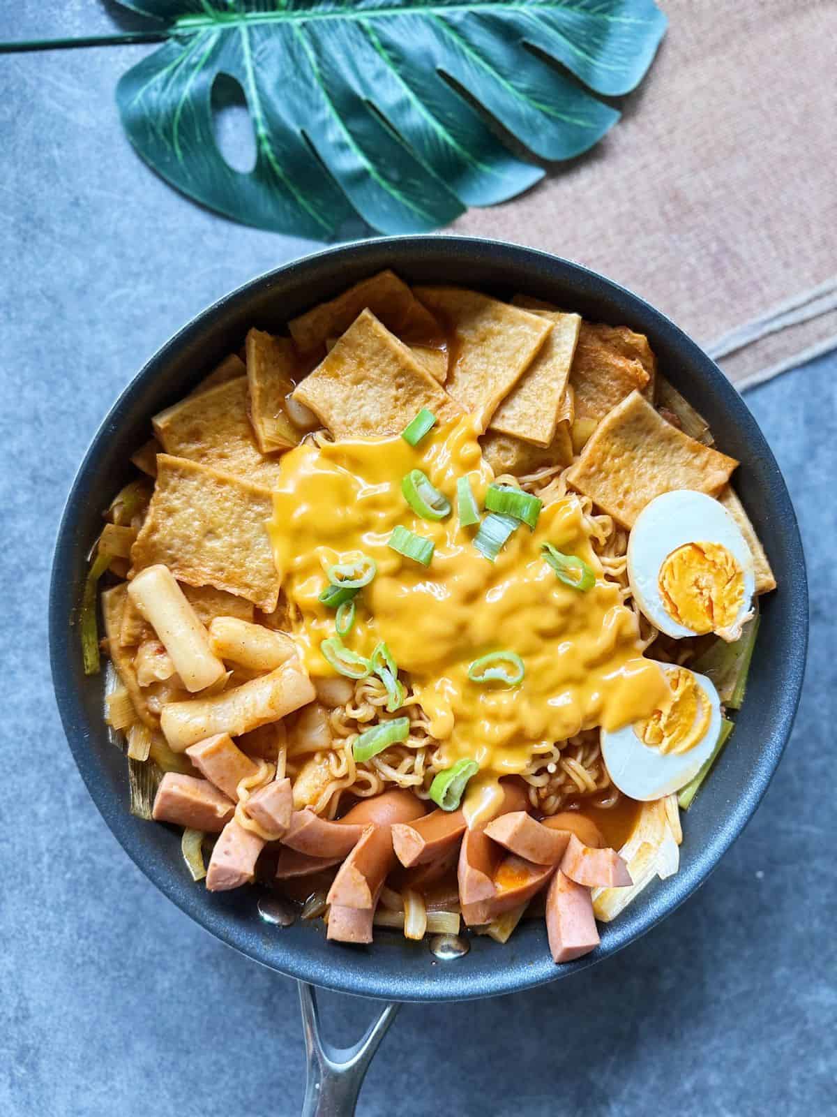 Pfanne mit würzigem Rabokki – koreanische Reiskuchen (Tteokbokki) und Ramyeon in roter Gochujang-Soße, mit Fischkuchen, Wurst, Ei, Lauch und geschmolzenem Käse garniert.