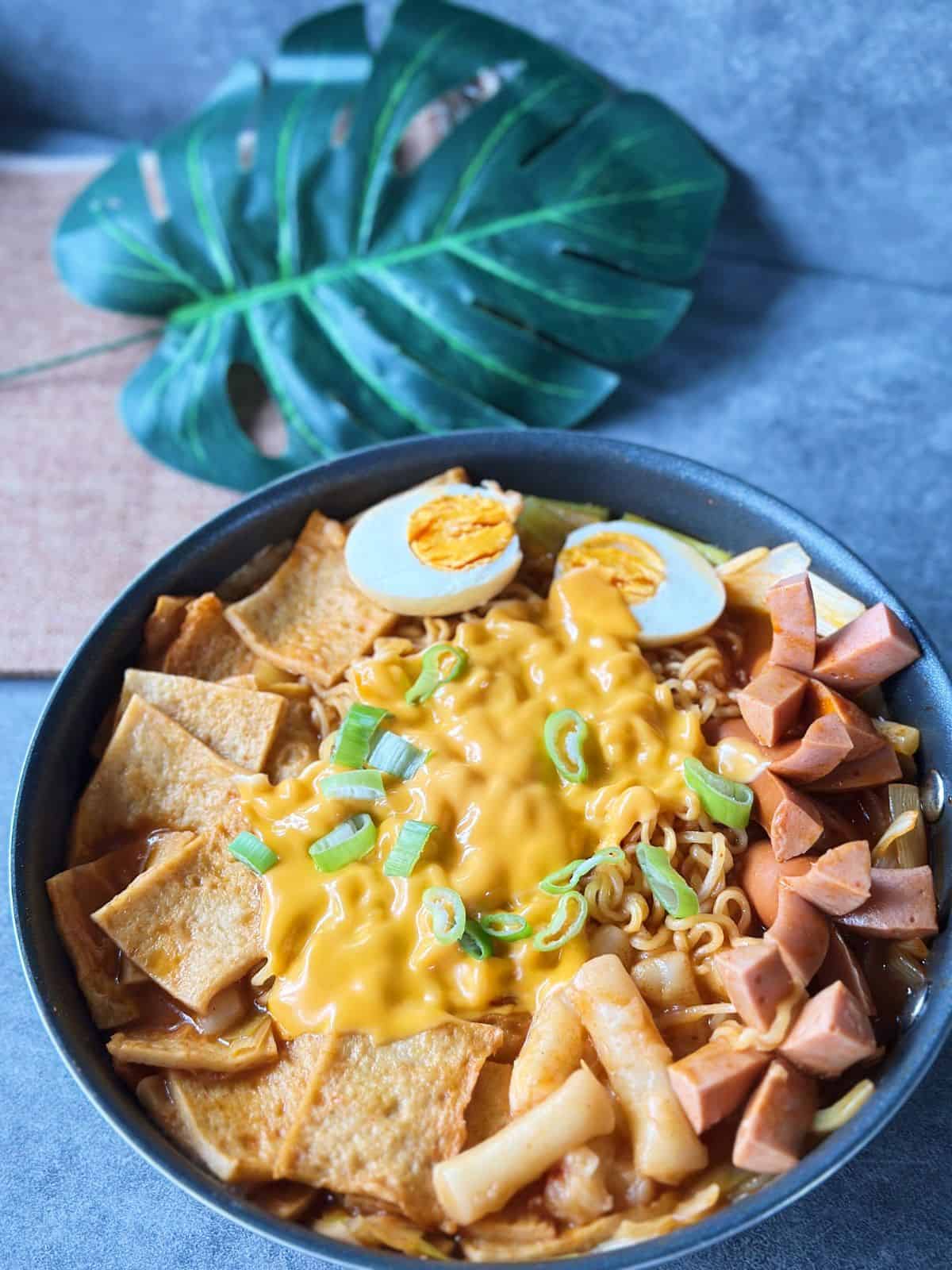 Pfanne mit würzigem Rabokki – koreanische Reiskuchen (Tteokbokki) und Ramyeon in roter Gochujang-Soße, mit Fischkuchen, Wurst, Ei, Lauch und geschmolzenem Käse garniert.