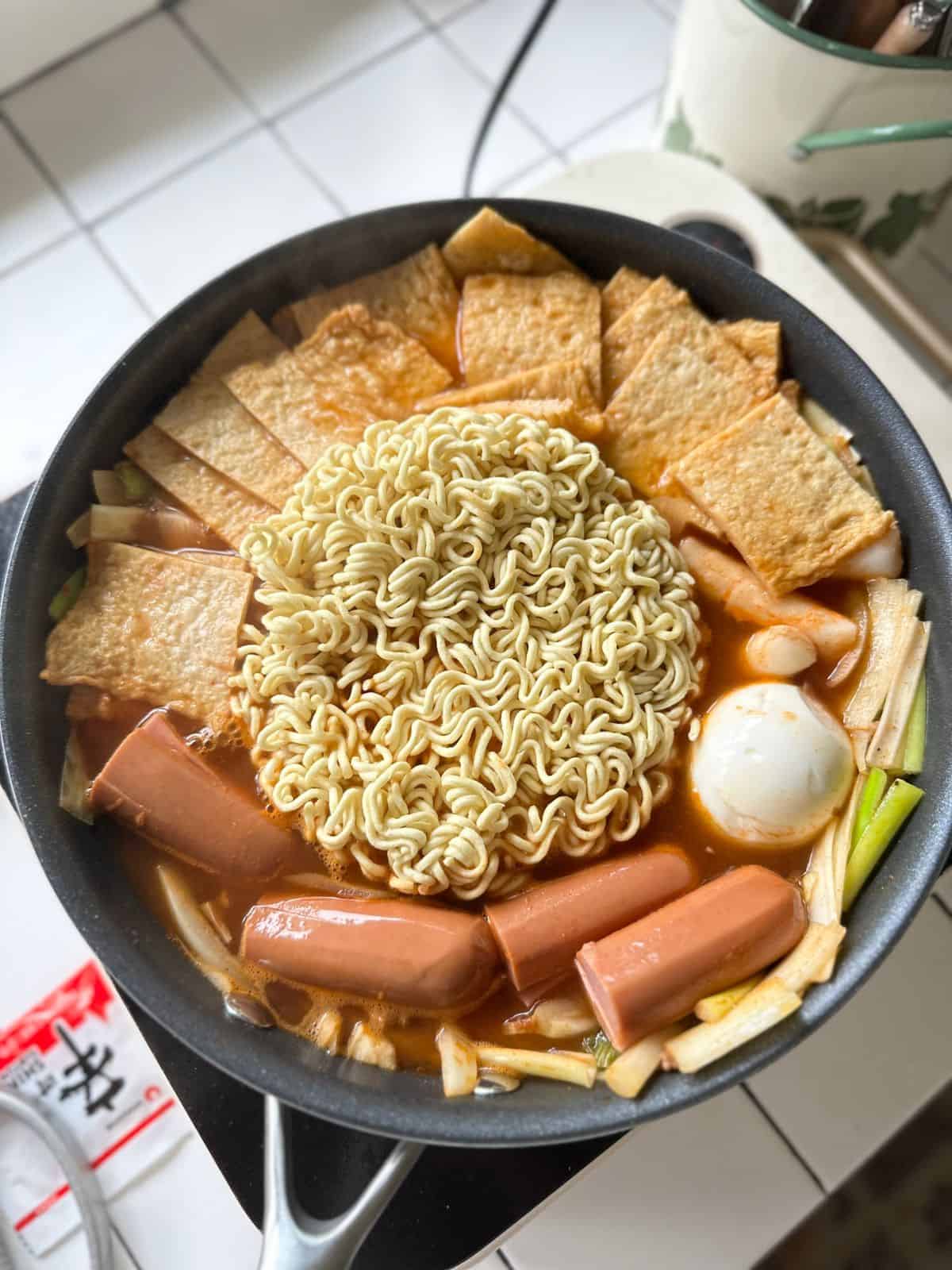 Pfanne mit köchelndem Rabokki – koreanische Reiskuchen (Tteokbokki), Ramennudeln, Fischkuchen, Wurstscheiben und hartgekochtem Ei in einer leuchtend roten, würzigen Gochujang-Soße.