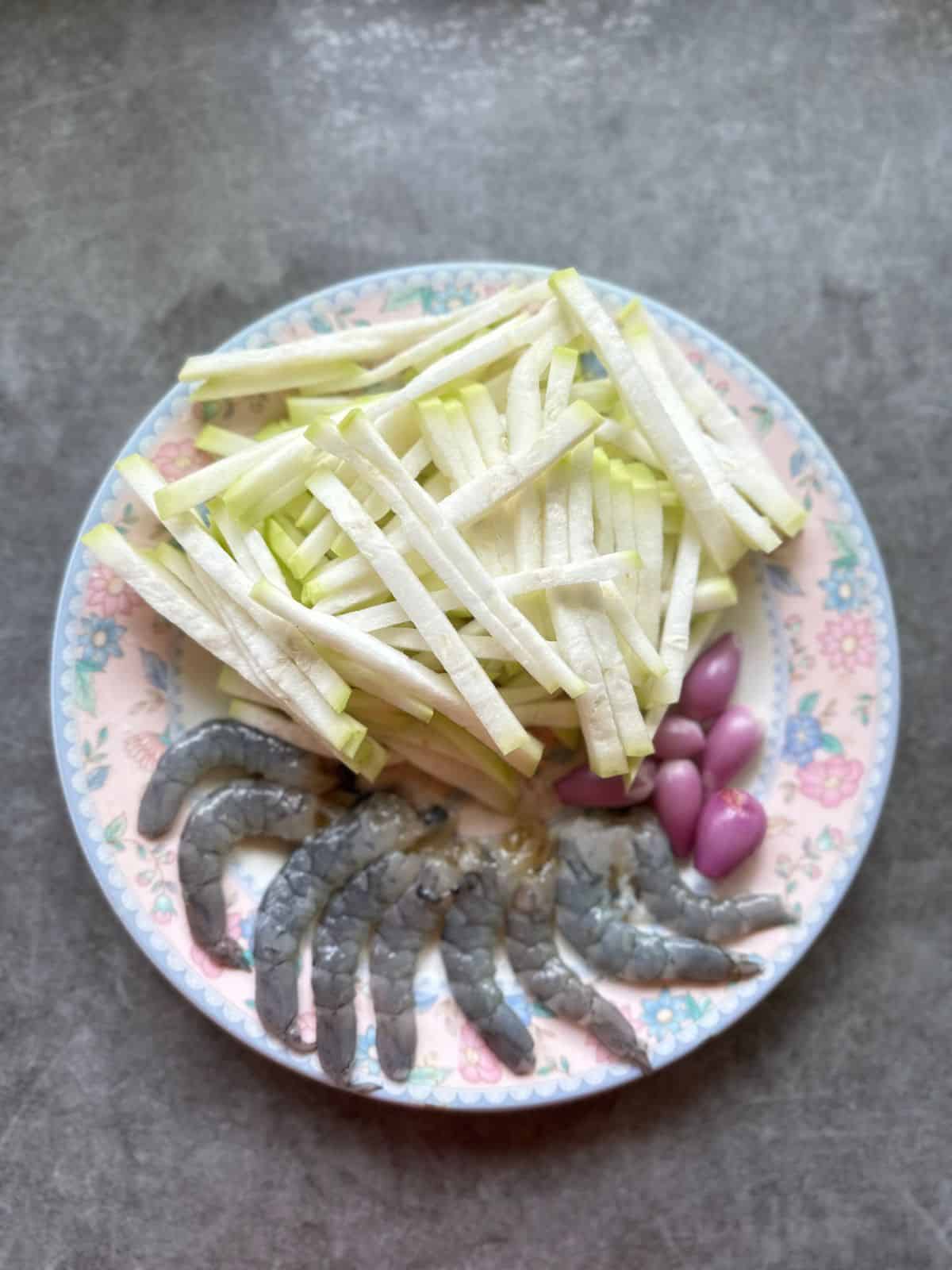 Thinly sliced opo squash, peeled shrimp with heads removed, and peeled shallots arranged on a plate, ready for cooking.