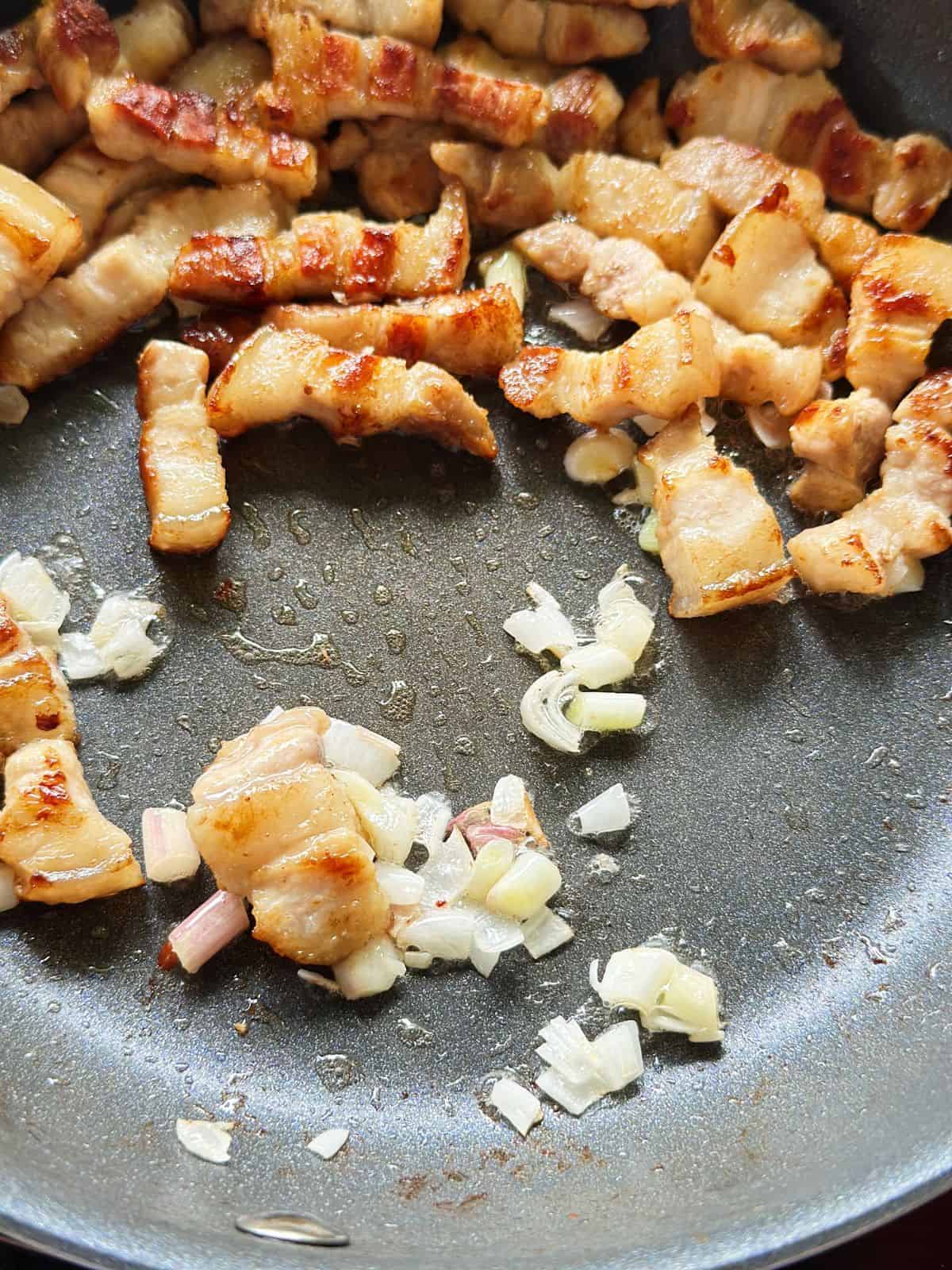 Stir-frying the white parts of scallions in a skillet after setting aside crispy pork belly pieces.