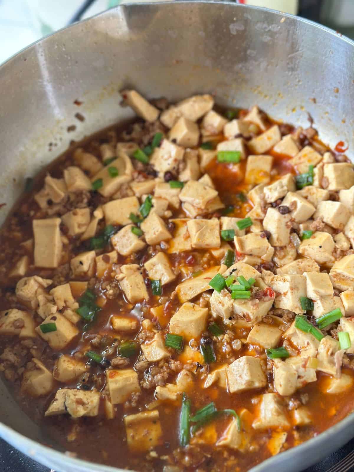 A pot of mapo tofu simmering in a rich red sauce, topped with freshly chopped scallions.